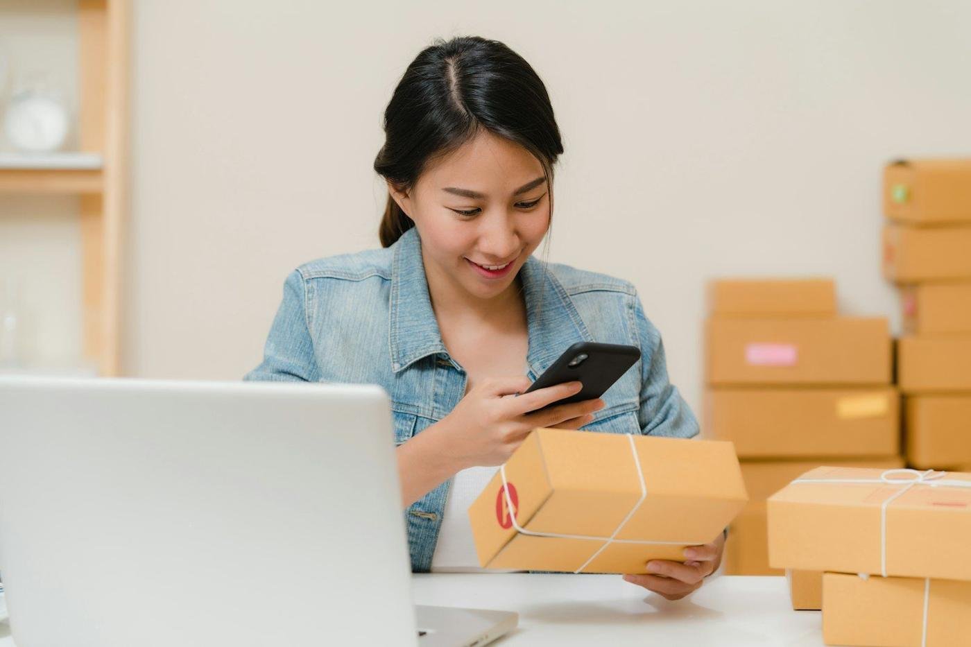 Woman checking a package with her phone near a laptop and boxes