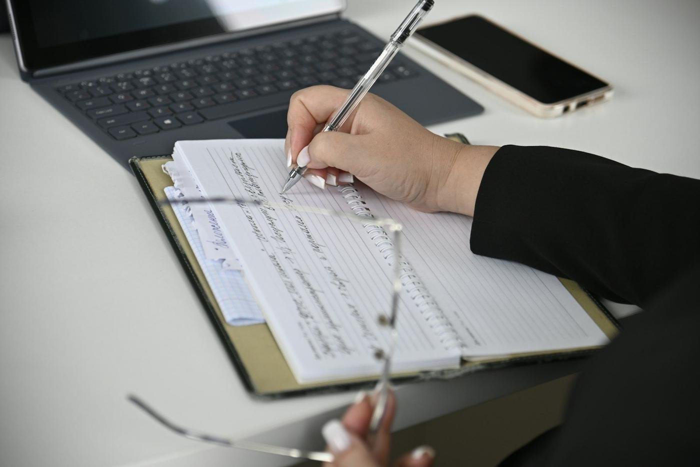 Person writing notes on a notepad beside a laptop and phone