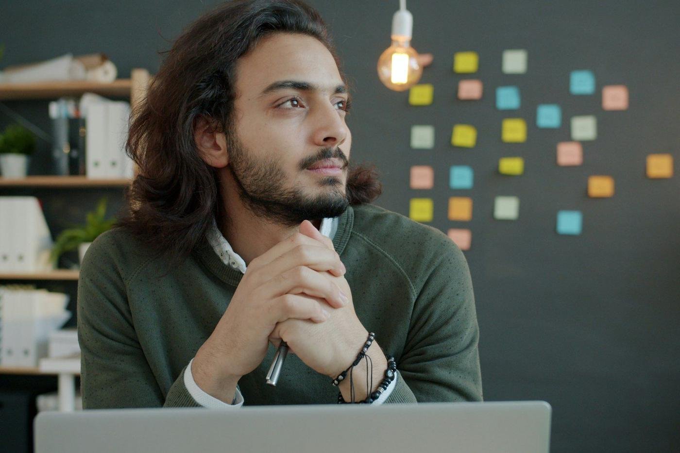 Man with long hair thinking at a desk with a laptop