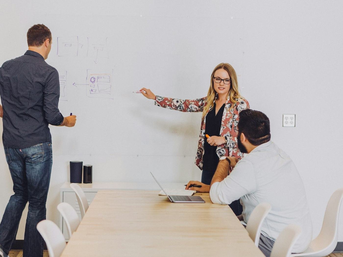 Small team meeting where a woman points at a whiteboard while colleagues stand and sit with a laptop
