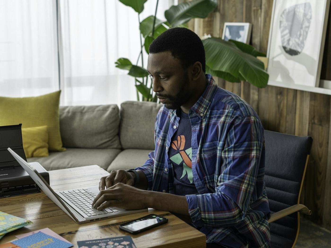 Man in a plaid shirt working on a laptop at a desk with a smartphone and notebook beside him