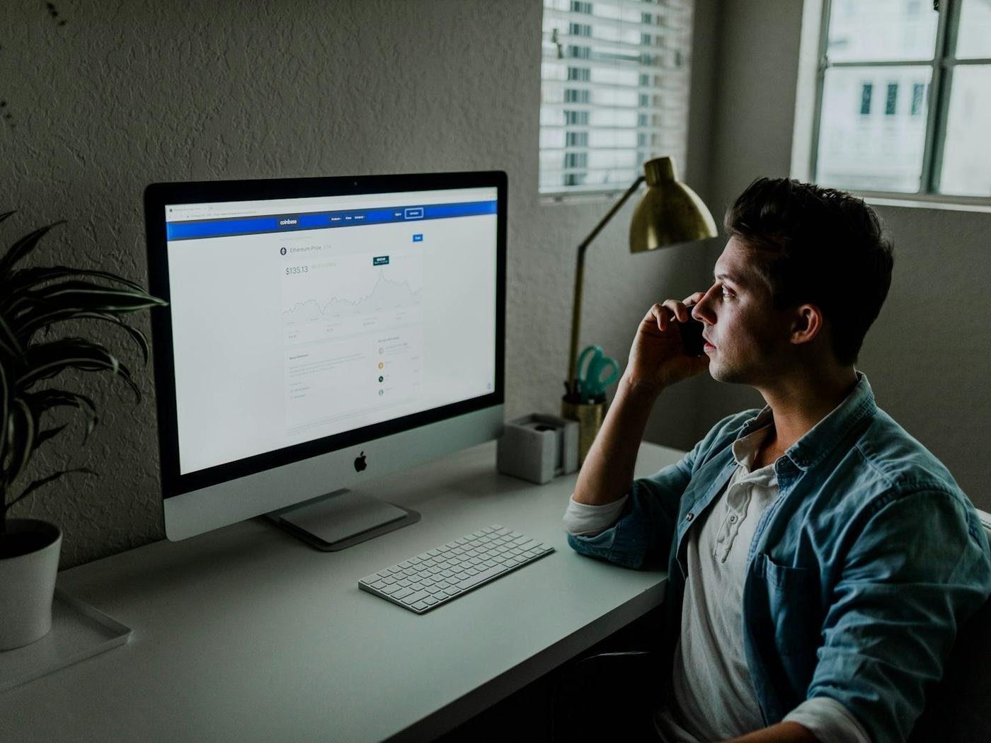 Man in a denim jacket at a desk facing a large monitor showing a web page